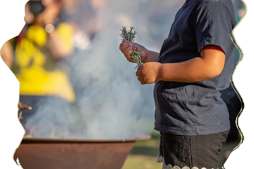 Un enfant tient de petites branches vertes au-dessus d'un grand feu d'où s'élève une fumée blanche.