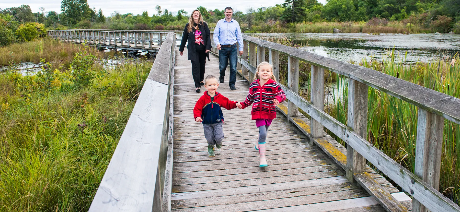 Un garçon et une fille courent en souriant sur une passerelle en bois au-dessus de l'eau, suivis de leurs parents.