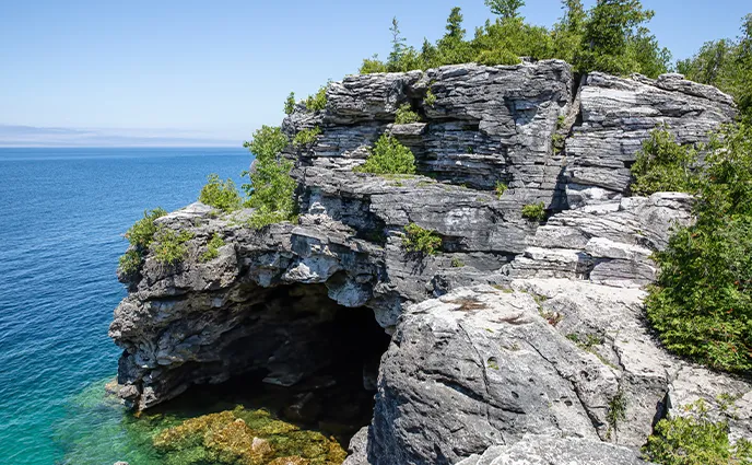 Une immense falaise de roche grise surmontée de sapins s'élève au-dessus d'une eau turquoise transparente.