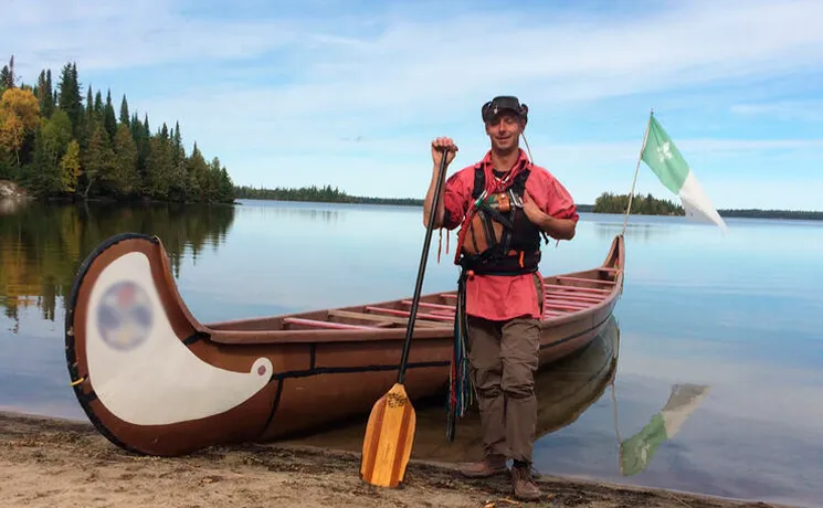 André Bernier debout avec une rame près d'un canot en écorce devant un lac et un drapeau franco-ontarien.