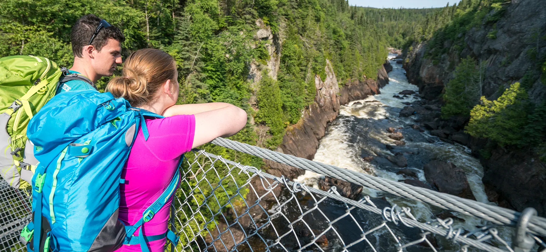 Un homme et une femme avec de grands sacs à dos regardent le paysage depuis un pont suspendu au-dessus d'une rivière.