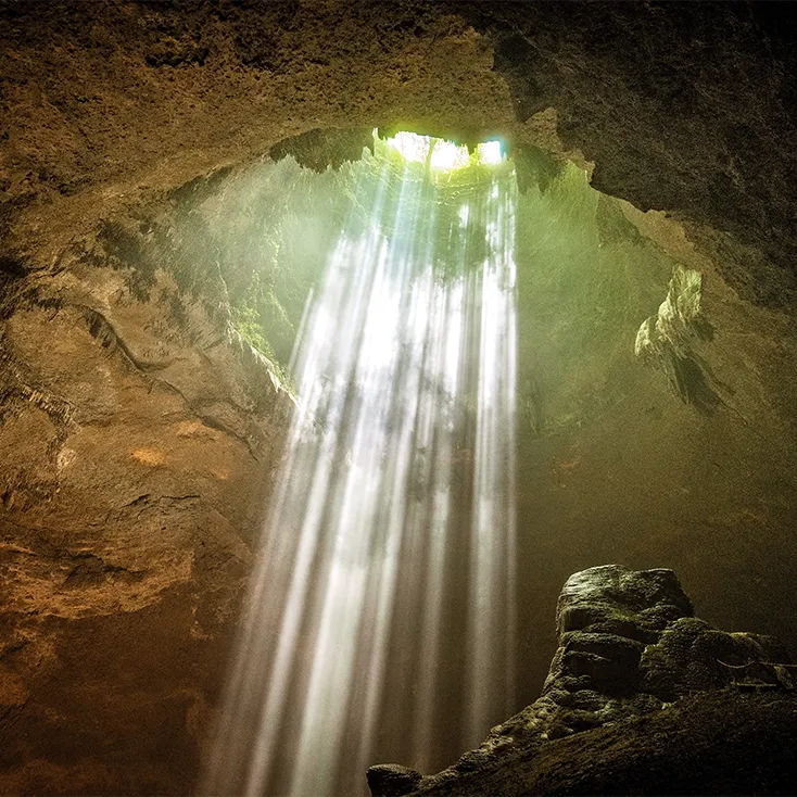 Photo de l’intérieur d’une grotte sombre où de grands rayons de soleil entrent par une ouverture au plafond.